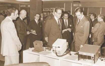 一群穿著西裝的男子在展覽會上圍著一個桌子上的電子設(shè)備觀看和討論。