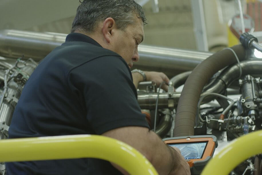 一位機械師在車間使用平板設(shè)備檢查機械，旁邊放置堅固工具箱和圓柱形工具，體現(xiàn)工業(yè)維修和技術(shù)的應(yīng)用。