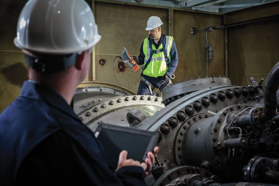 一名工人穿著安全帽和制服，使用堅固平板設(shè)備在工業(yè)車間檢查重型機械，背景有堅固工具箱和工廠環(huán)境，顯示數(shù)據(jù)分析和監(jiān)控過程。