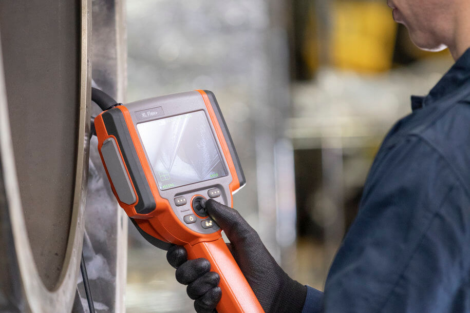 一名技術(shù)工人穿著安全手套和制服，使用手持式檢查設(shè)備和攝像頭探頭在工廠車間進行機械維護和檢測，屏幕顯示實時監(jiān)控畫面。